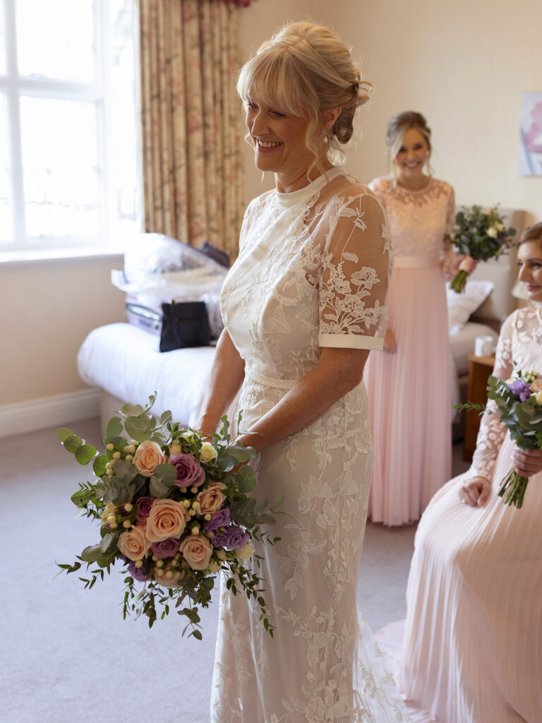 Bride stood looking at herself in the mirror. She is holding a bouquet of pale pink, purple and ivory roses.