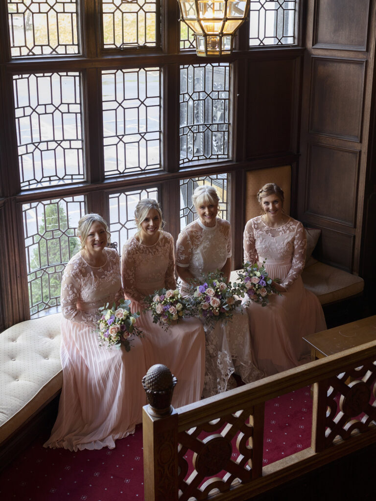Bride and bridesmaids sat down in a seating area on the stairs at The Petwood Hotel. There are big windows behind them.