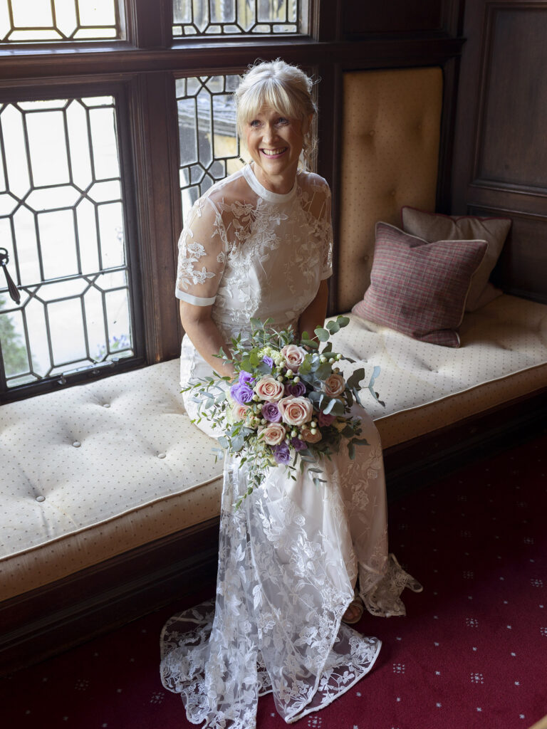 Bride sat down in a seating area on the stairs at The Petwood Hotel. There are big windows behind her. She is looking to the side at her Bridesmaids who are out of the shot