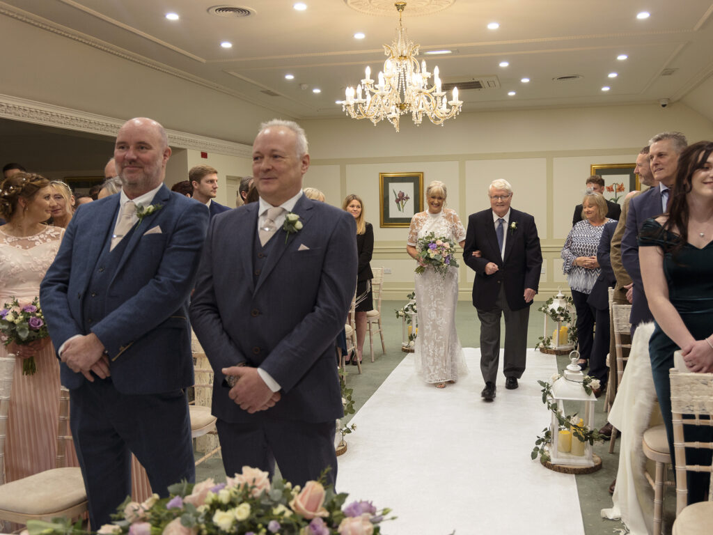 A Groom and his best man are stood at the top of the aisle with his Bride and her father walking up the aisle towards them