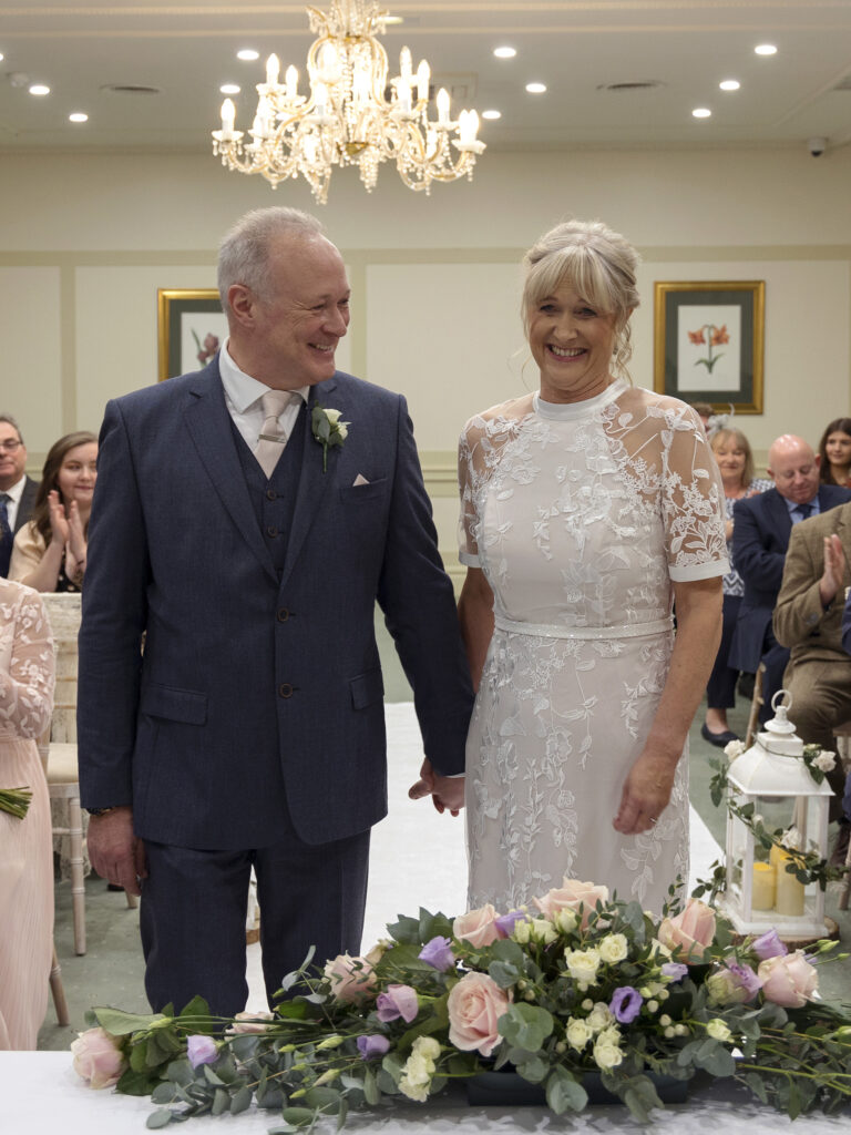 A Bride and Groom are stood at the top of the aisle holding hands. She is smiling and he is looking at her smiling too