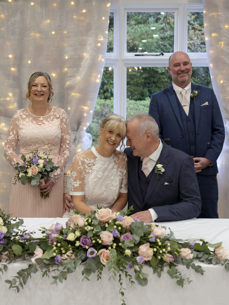 Bride and groom are sat at a table after signing the wedding register. He is lenaing his forehaed agaianst hehr face. There is a bridesmaid and best man stood behind them smiling at the camera