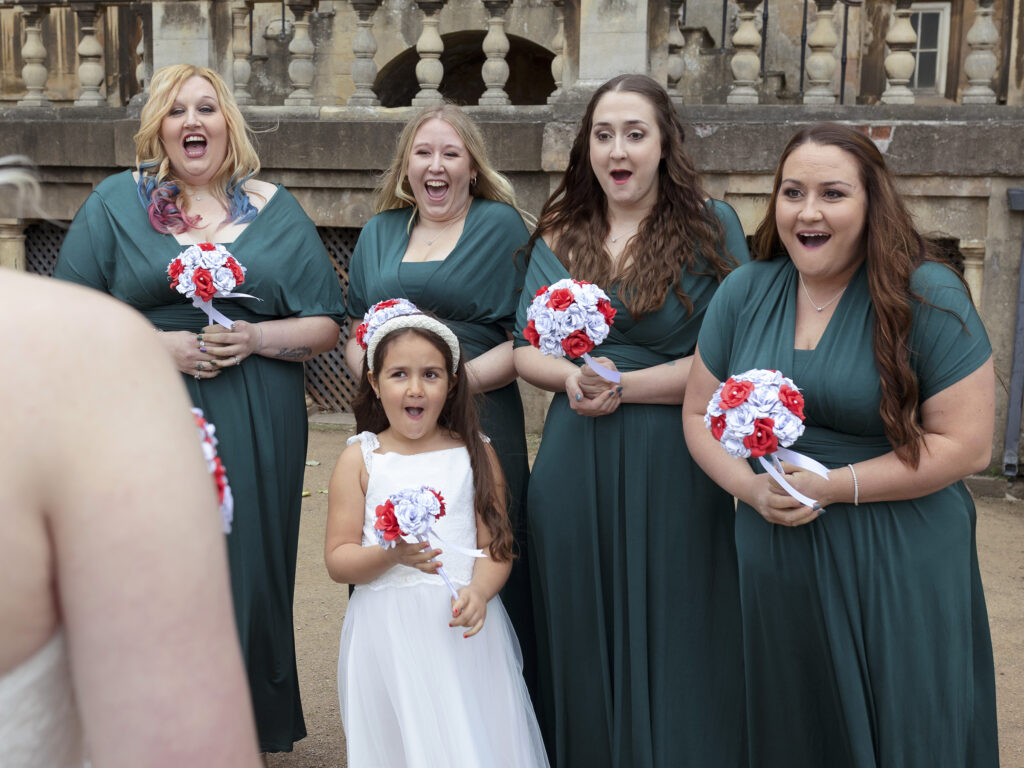Bridesmaids stood in a row, seeing the bride in her dress for the first time. They all have happy, shocked and emotional expressions