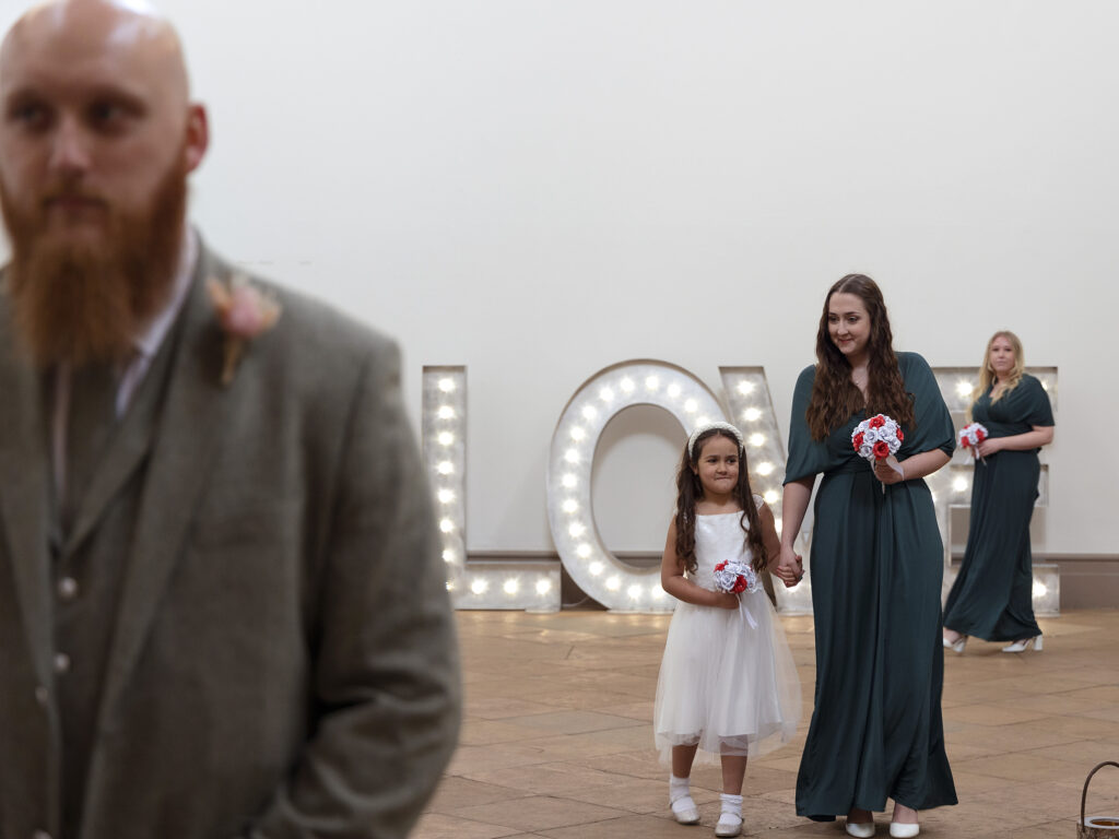 Groom is stood close to the camera at the front of the aisle and bridesmaids are walking towards him at Wollaton Hall