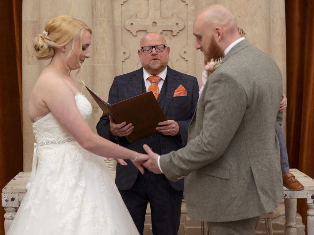 A bride and groom are stood holding hands at the top of the aisle. The celebrant is conducting the wedding ceremony at Wollaton Hall
