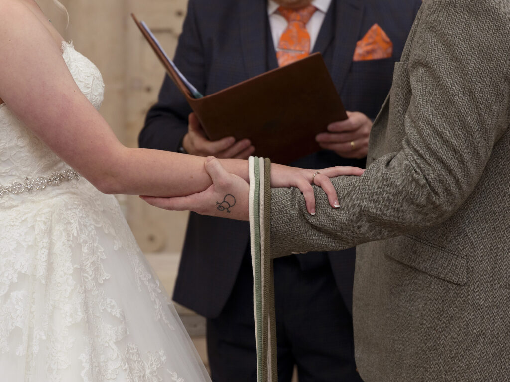 A bride and groom are holding arms and have a ribbon laid over the top. The celebrants book is in the background at Wollaton Hall