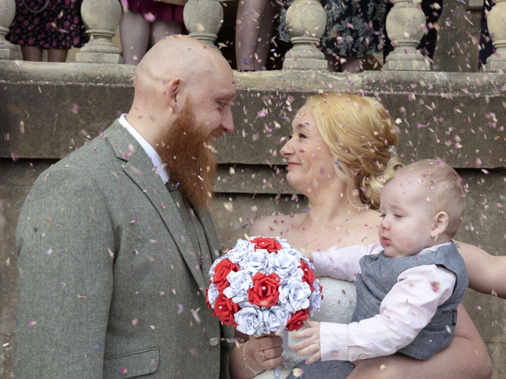 Close up of Bride and groom with their son, guests are throwing confetti over them outside Wollaton Hall
