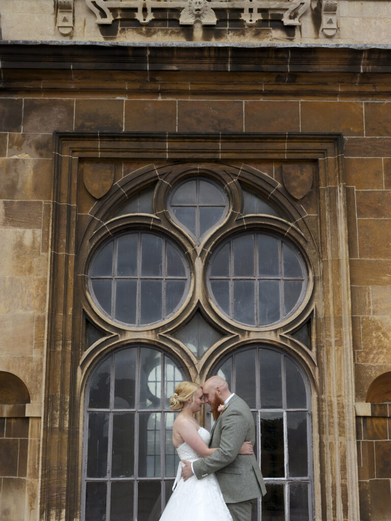Rooftop of Wollaton Hall. The bride and groom are hugging and have their faces close to each others. Mid tight crop. The couple are stood in front of a large ornate window