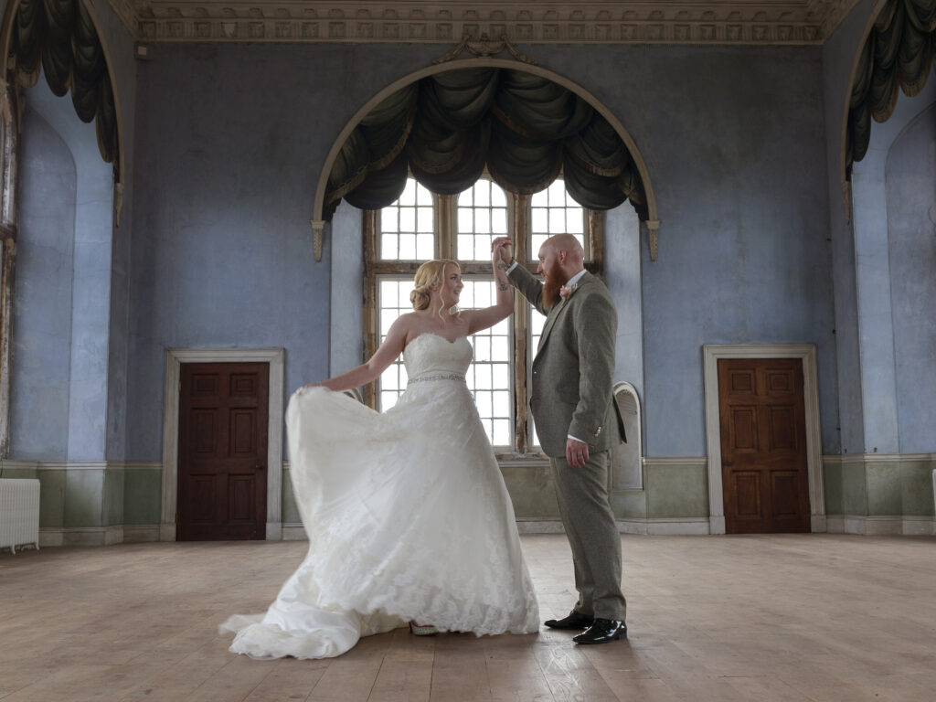 A bride and groom are stood facing each other in the Blue Prospect room in Wollaton Hall. The Bride is swishing her dress around