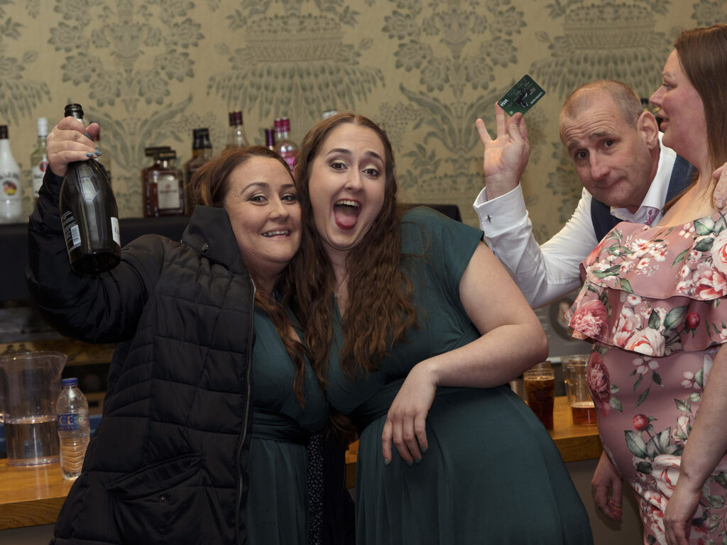 guests at a wedding at Wollaton Hall. 2 bridesmaids are smiling for the camera with a bottle of prosecco. Their dad is in the background waving his credit card in the air.