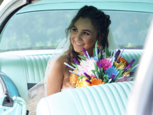 Colourful Summer wedding. A bride is sitting in the back of a classic car. shes looking out the window smiling over the top of her flowers