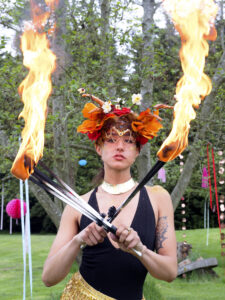 Colourful Summer wedding. A fire performer stands looking directly at the camera. She is holding fire sticks is a cross and is wearing a floral headdress