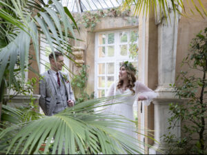 Modern Greenery Wedding. Bride and groom are standing facing each other, leaning on the walls of the conservatory at Harlaxton manor