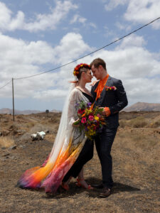 Lanzarote Elopement. Bride and Groom are standing together in the Lanzarote lanscape. She is wearing a brightly coloured dip dye dress and he has an orange shirt on with a traditional suit.