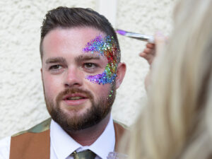 Real wedding of two grooms. A Groom is sat having his face glittered in pride rainbow colours