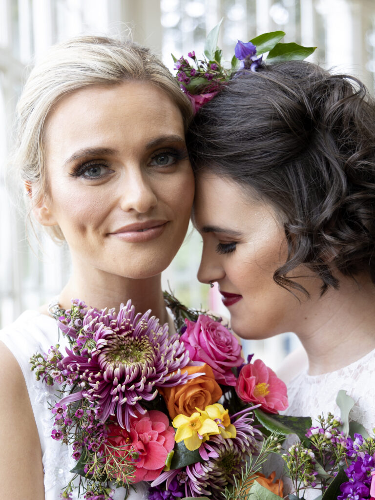 Two brides with their faces close to each other. Tightly cropped photo of their faces. Blonde Bride looks at the camera, while the dark haired bride rests her forehead on her wifes cheek and closes her eyes.
Both have brightly coloured flowers as a necklace and hair piece.
