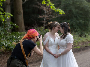 Brightly colour haired Photographer is taking photos of 2 Brides on their wedding day. They are leaning their foreheads together and holding hands in front of them. They are stood in the woodland of Clumber Park