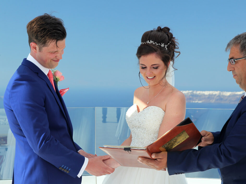 bride and groom reading vows with the Santorini sea behind them. The groom is wearing a blue suit and the bride has a big princess style dress.