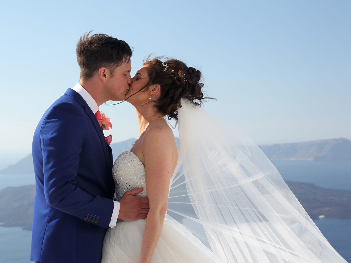 Bride and groom share a romantic kiss during a destination wedding portrait in Santorini with ocean and cliffs in the background.