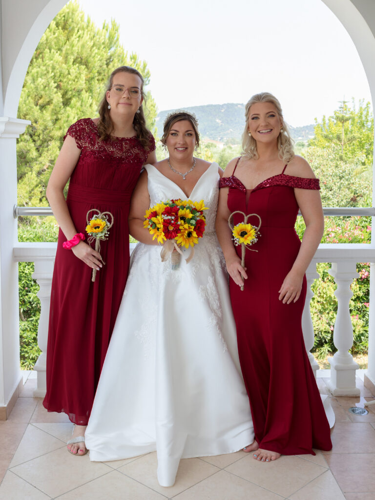 Bride stood with a bridesmaid either side. The bridesmaids are wearing deep red dresses and they are all holding sunflowers