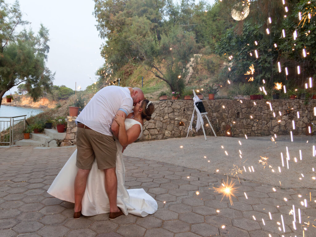 Bride and groom are having their first dance in Zante. He is dipping her and giving her a kiss. there are sparklers/fireworks going off on the right hand side.