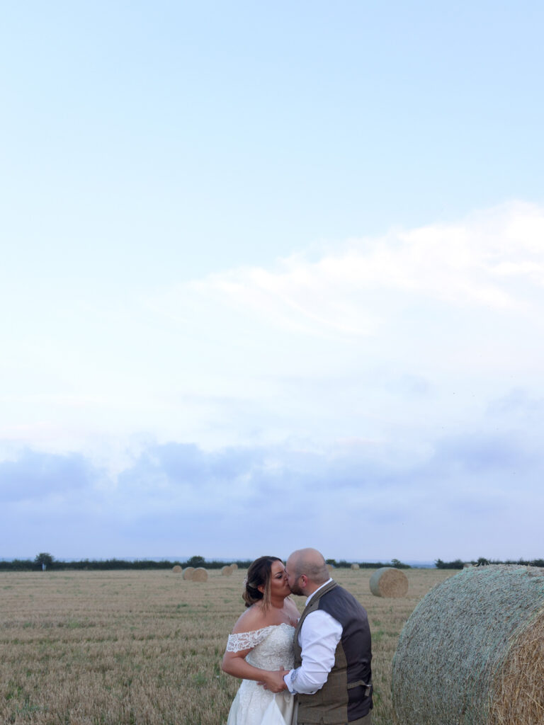 Bride and groom in the centre bottom of the photo kissing. They are stood in a wheat field. The majority of the photo is made up of blue sky