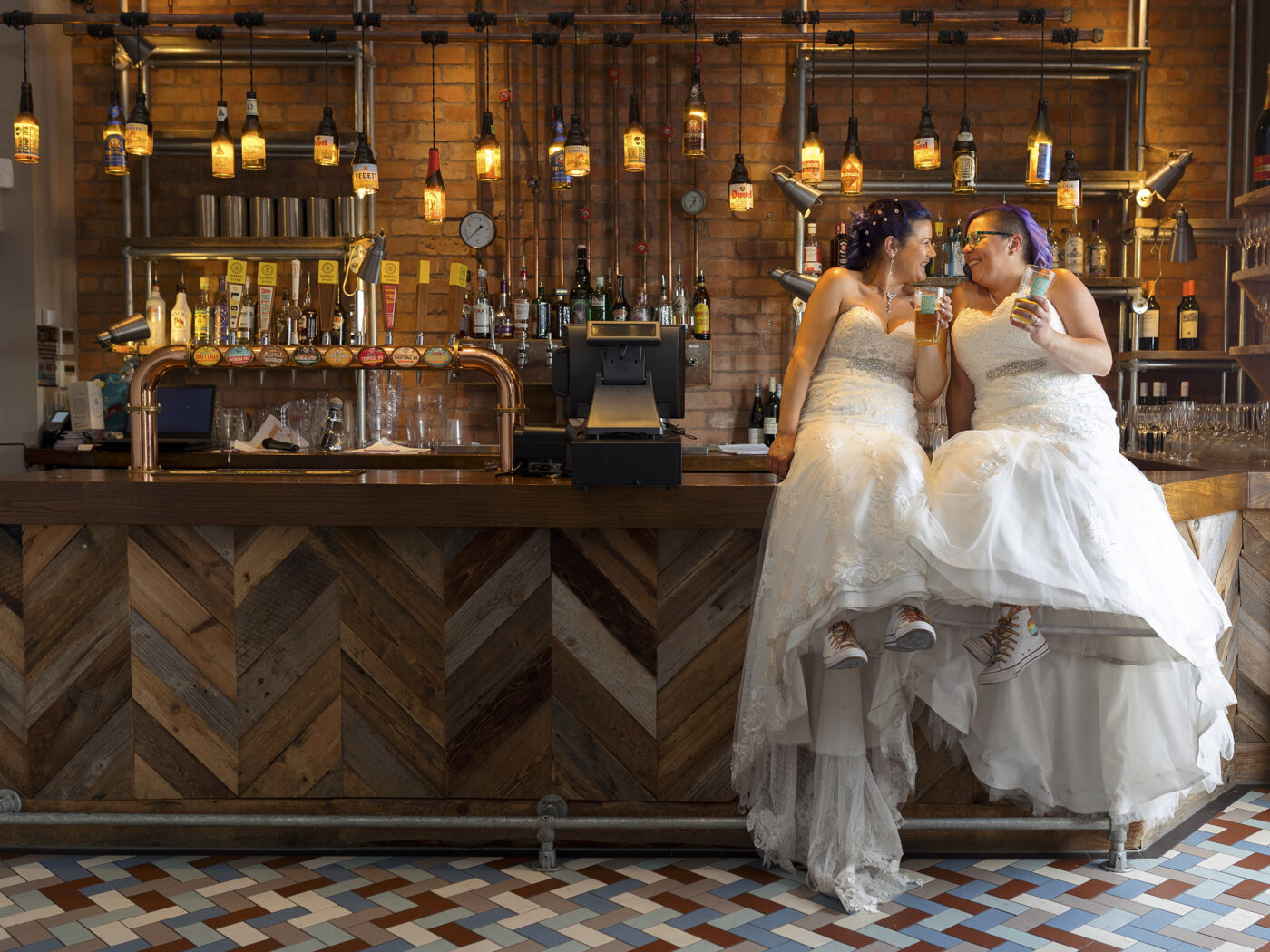 Nottingham wedding blog wo brides in wedding dresses sit at a rustic bar clinking drinks together in a warm, stylish wedding reception setting.