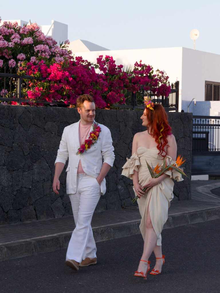 Bride and groom walking side by side along the road in Lanzarote . The groom has a bright floral lapel on his white suit and there is bright pink bougainvillea on the volcanic stone wall behind