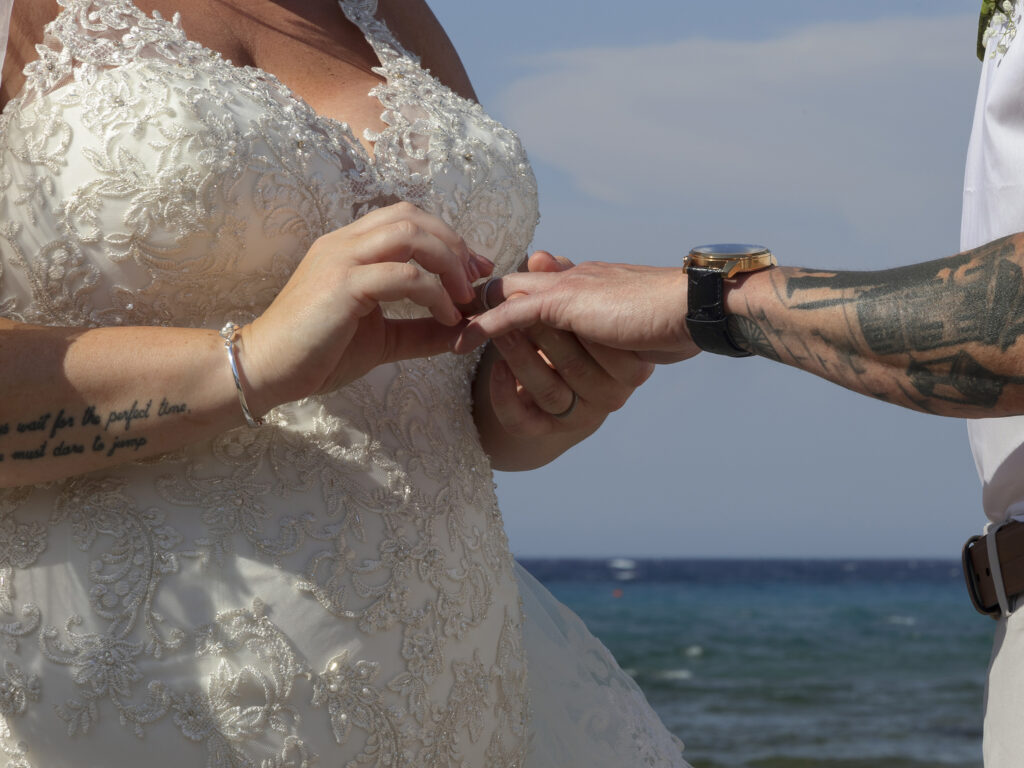Close up of rings being exchanged. The bride is putting a ring on her grooms finger. the sea is in the background