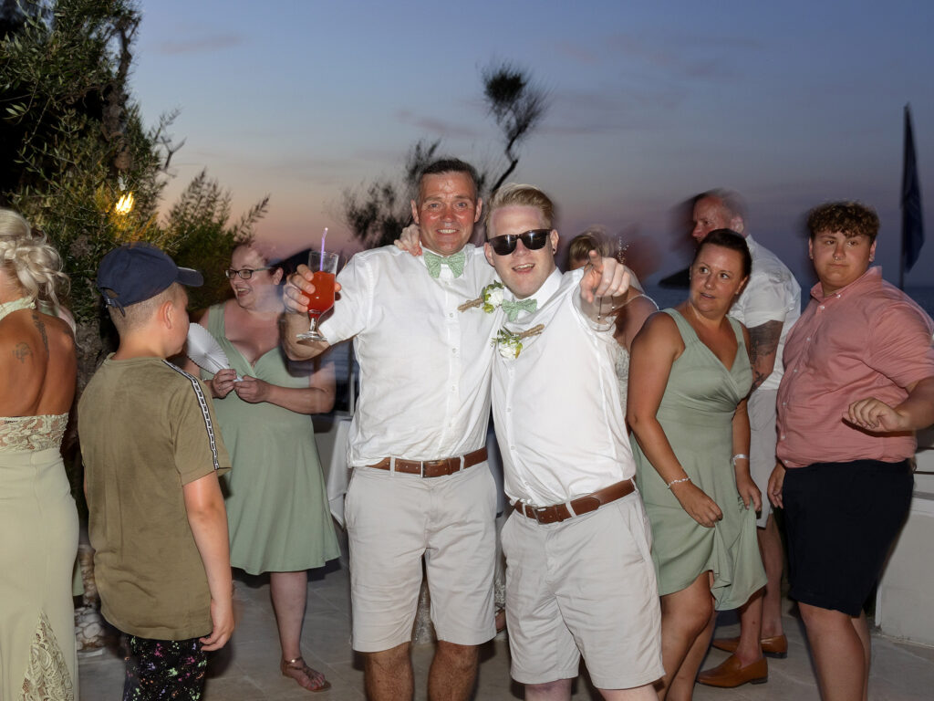 Guests of a destination wedding are dancing on the dance floor. The Zante sun is setting and 2 guests are hugging and pointing towards the camera
