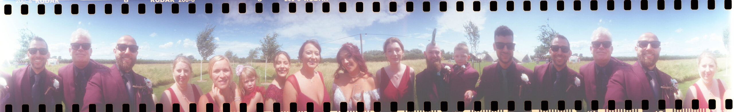 Panoramic photo of a bride and groom with their bridal party. The photo is a lomography 360 photo so has the camera sprocket holes and film numbering.