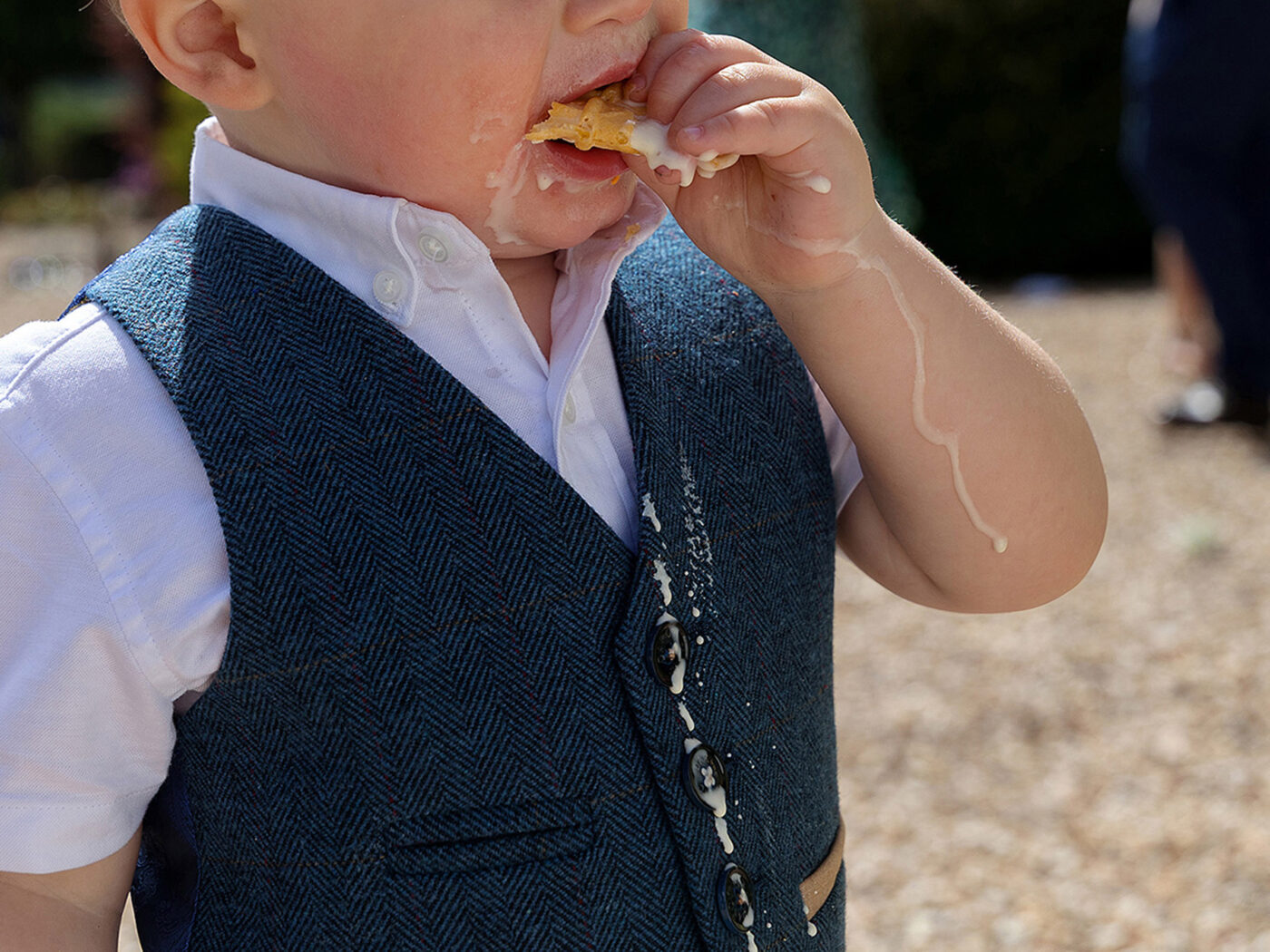 Close-up of a young child in a blue waistcoat eating an ice cream cone, melted ice cream dripping down their hand and vest.
