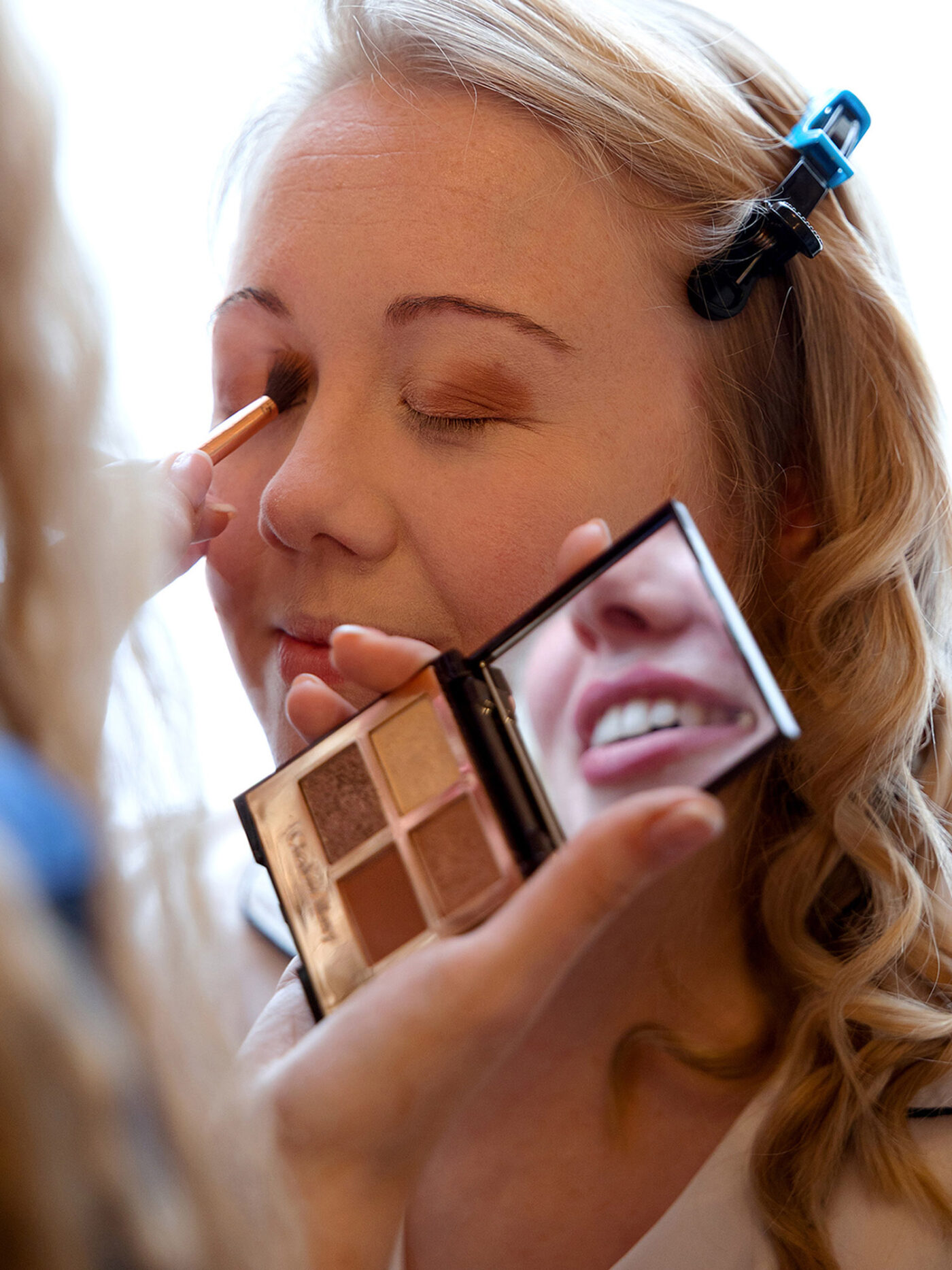 Close-up of a woman having brown eyeshadow applied while the MUA is holding an open makeup palette with a mirror.