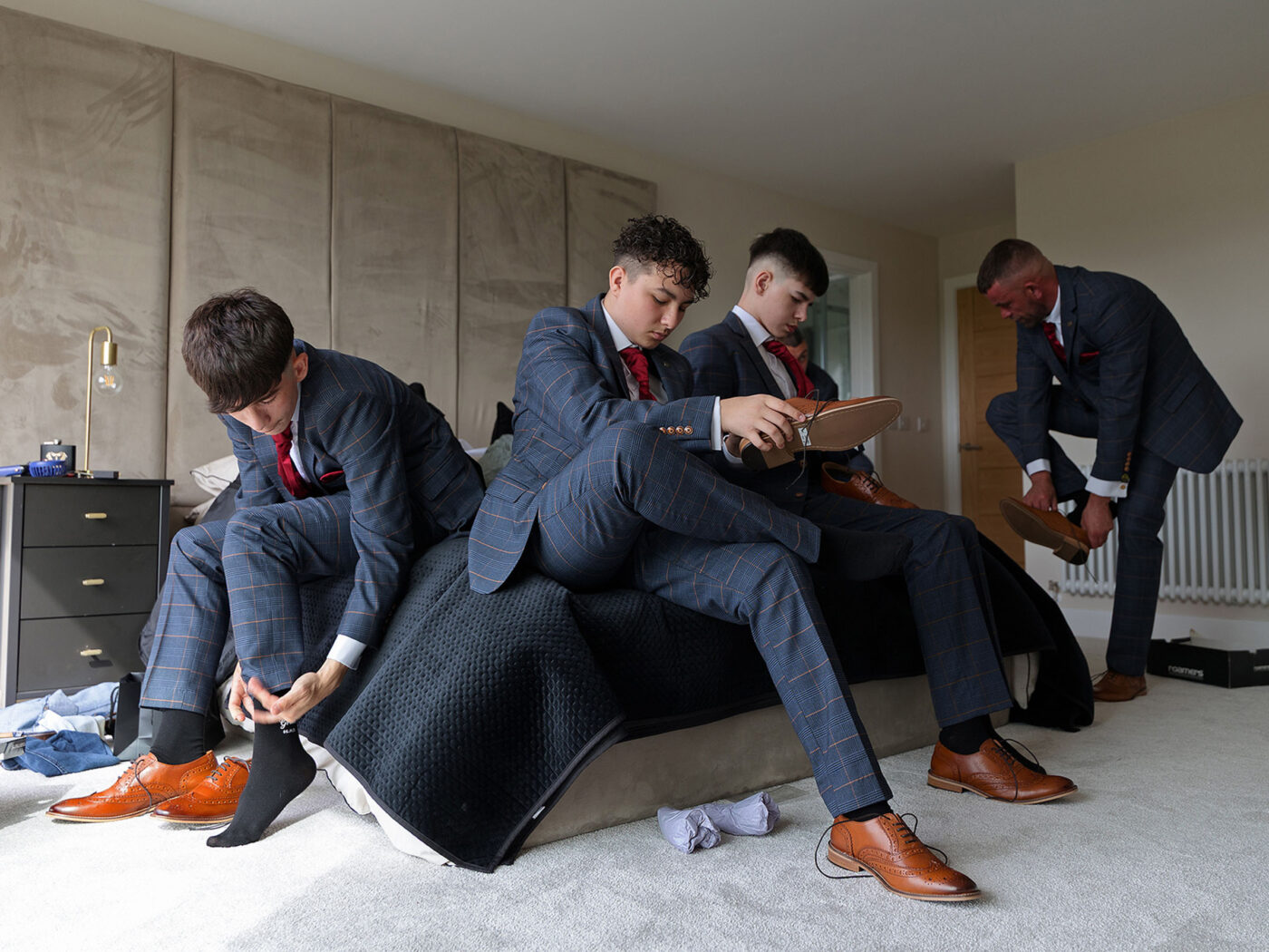 Three young men in matching blue suits with red ties sit on a bed putting on brown dress shoes in a modern bedroom.
