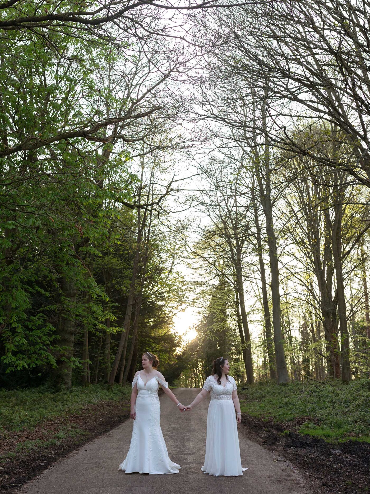 Two brides in white gowns stand holding hands on a tree-lined path at sunset, facing opposite directions.