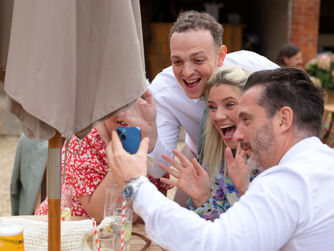 A group of guests crowd around a smartphone at a table, reacting excitedly and laughing together.