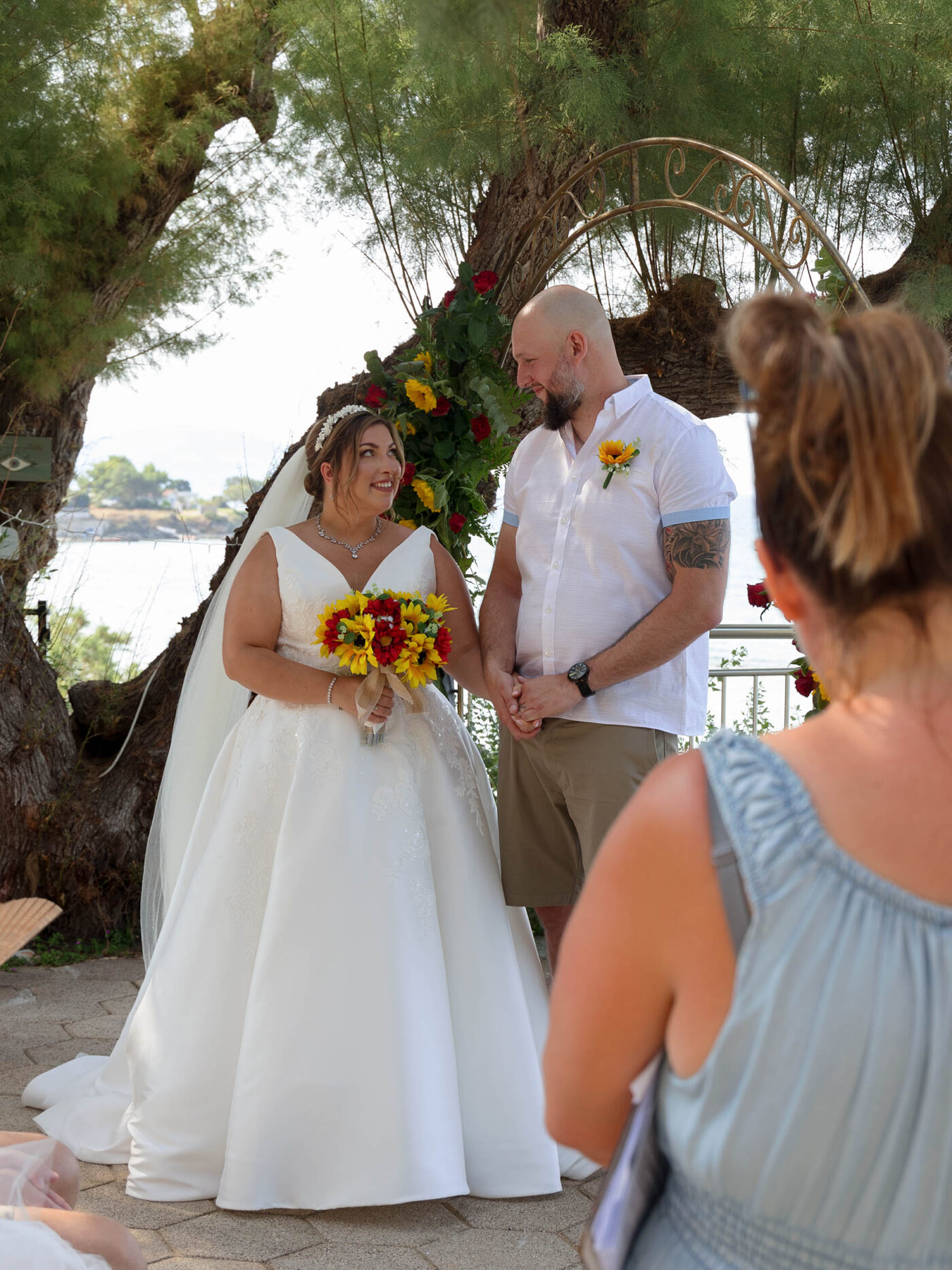 A bride and groom stand outdoors in Zante by the sea during their ceremony, holding hands beneath a circular floral arch while a guest looks on.
