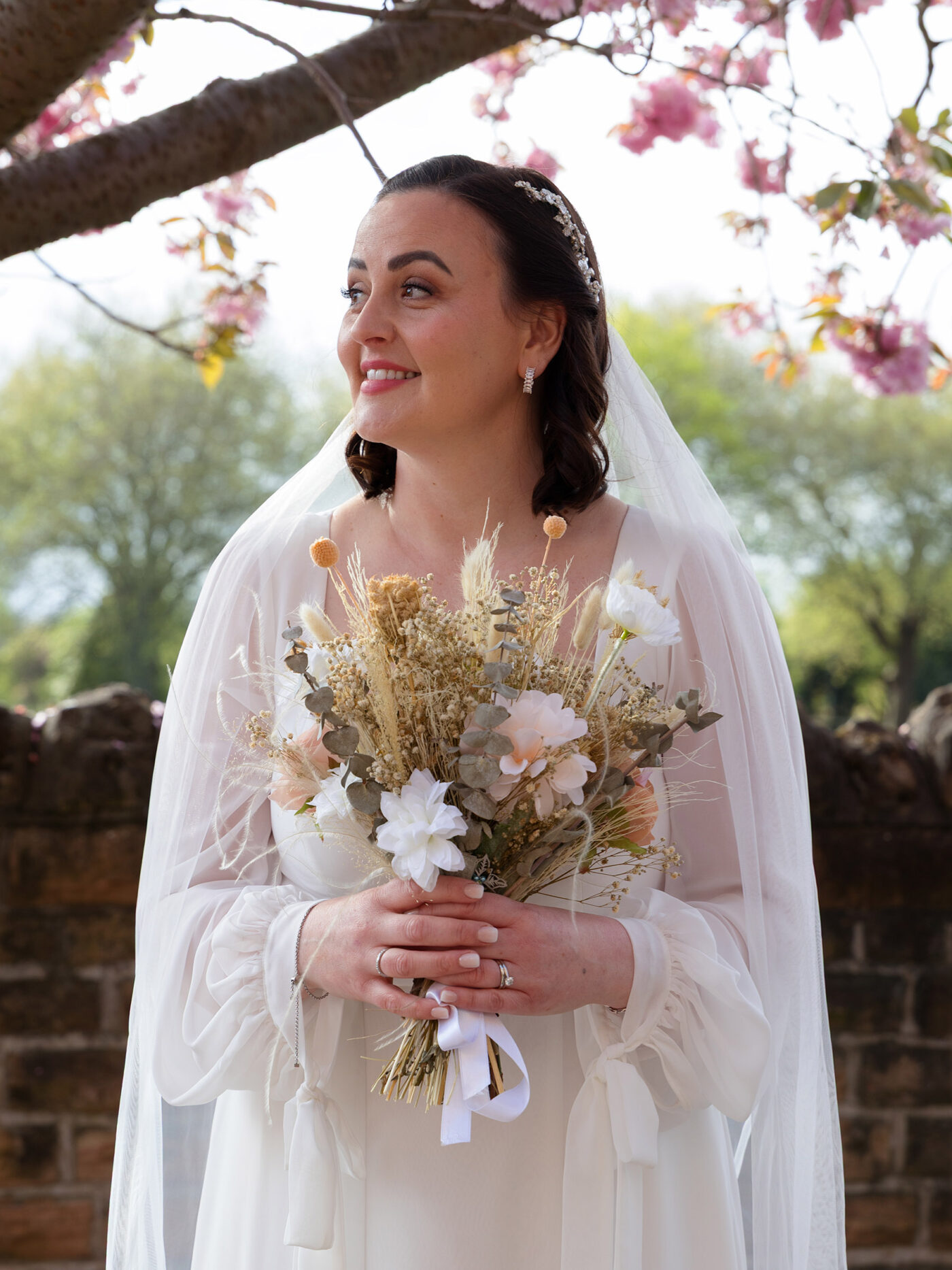 A bride in a white gown and veil holds a bouquet of dried and pastel flowers, standing beneath blooming pink blossoms.