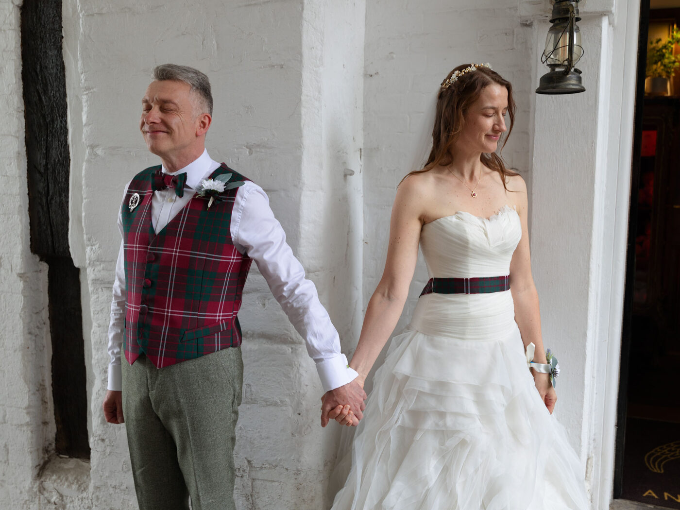 A Bride and groom stand holding hands around a corner to each other. She is waering a wite wedding dress and he has on a tartan waistcoat