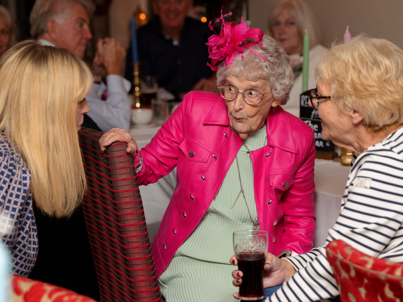 An elderly woman in a bright pink outfit and fascinator chats animatedly with two seated guests at a reception table.