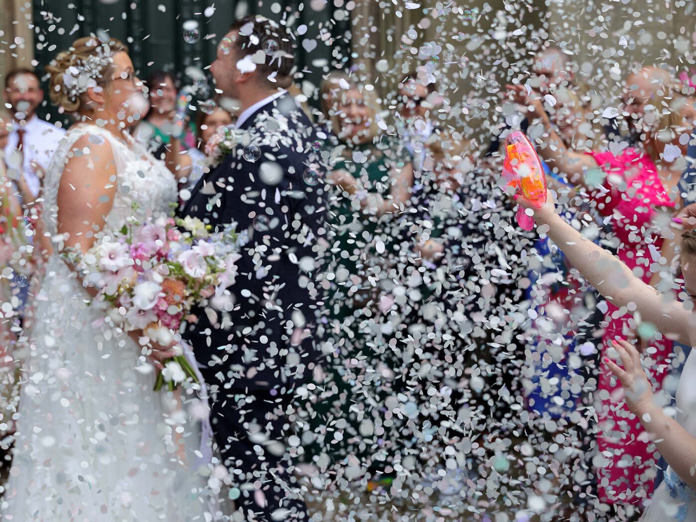 Bride and groom laugh as colourful confetti is sprayed over them during an outdoor celebration.