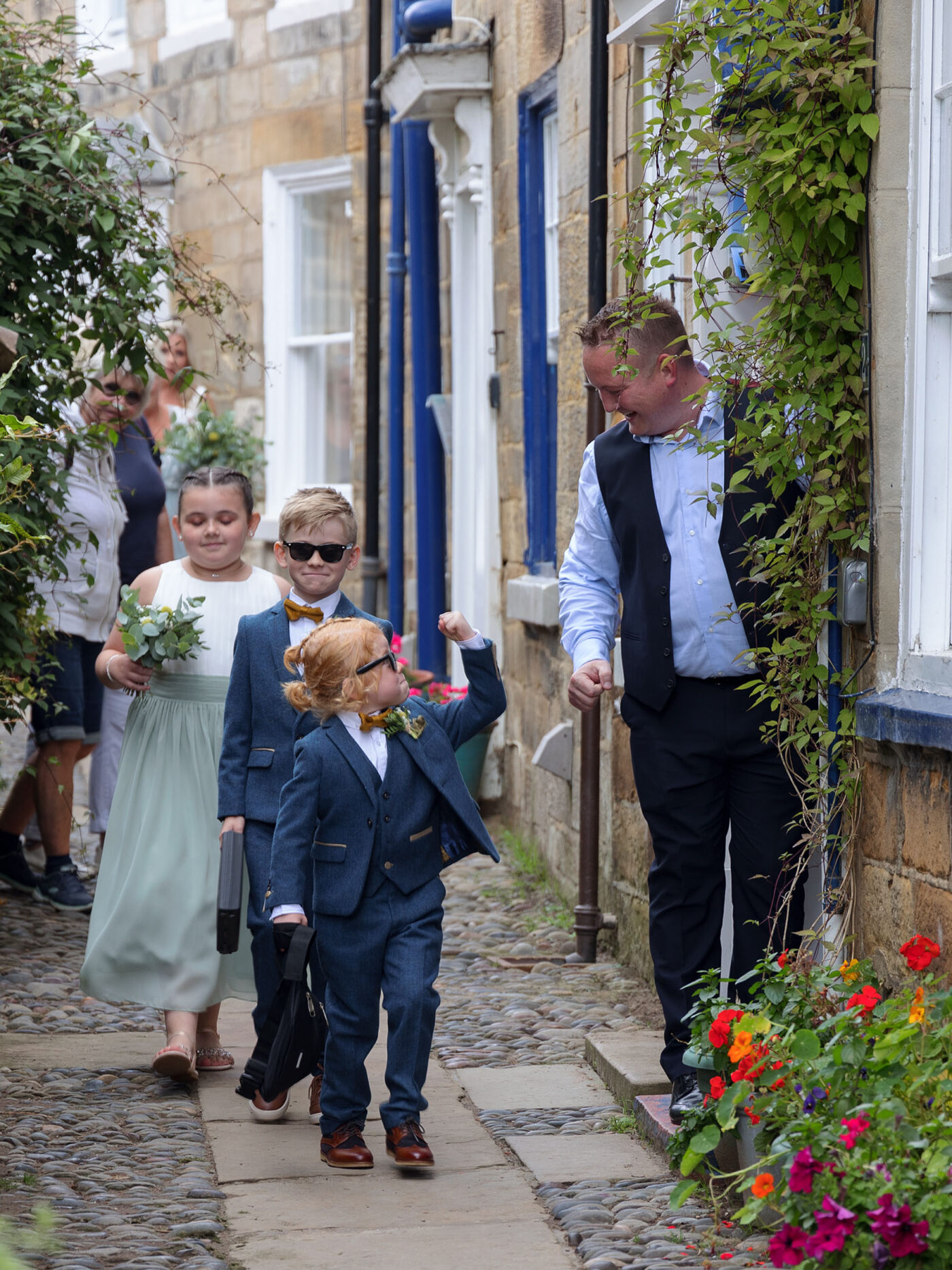 A young page boy in a blue suit walks confidently down a narrow cobbled street, followed by bridesmaids and a smiling adult.