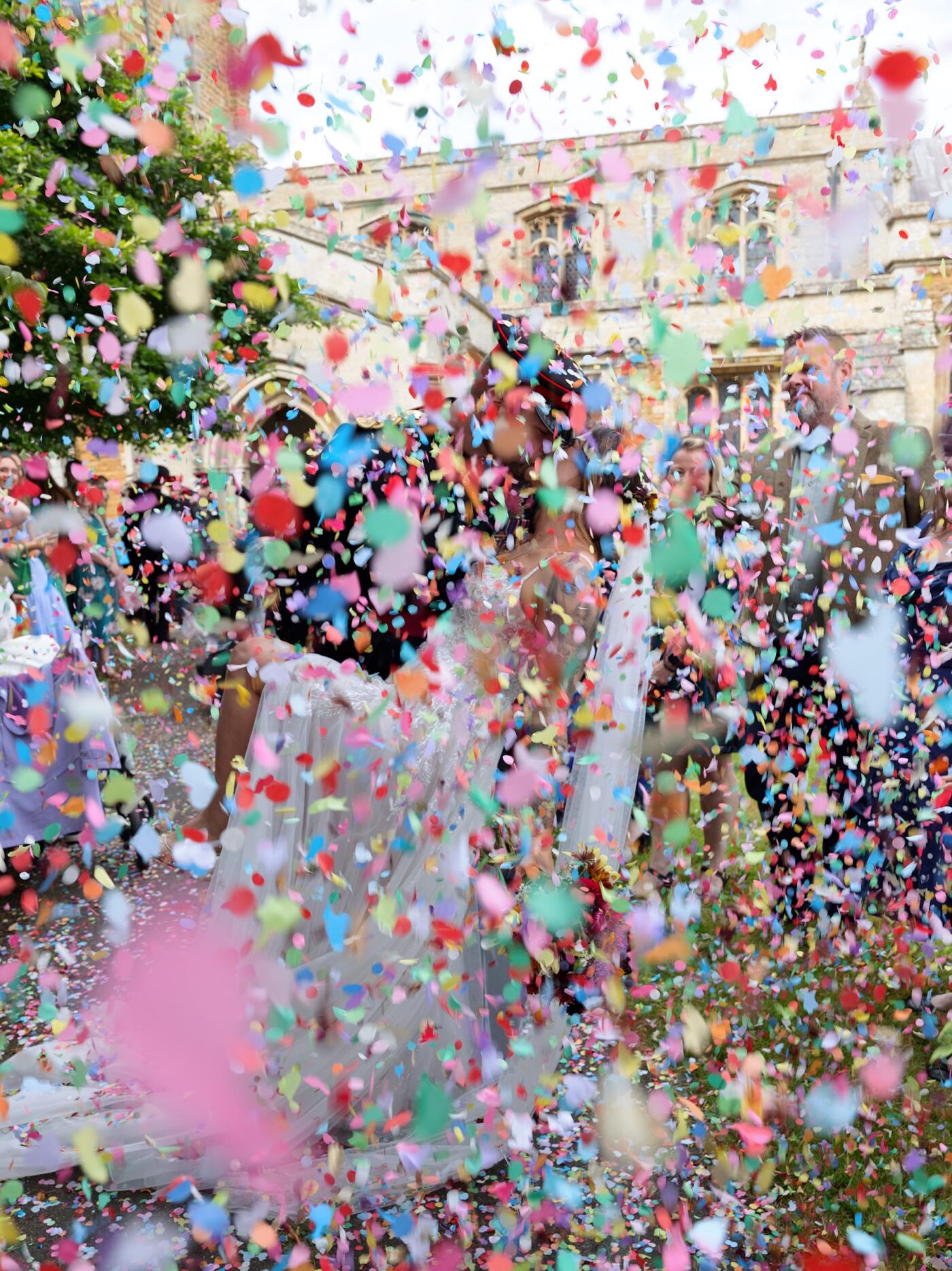 A joyful outdoor confetti moment with guests celebrating as the couple stand together beneath a shower of colourful paper.