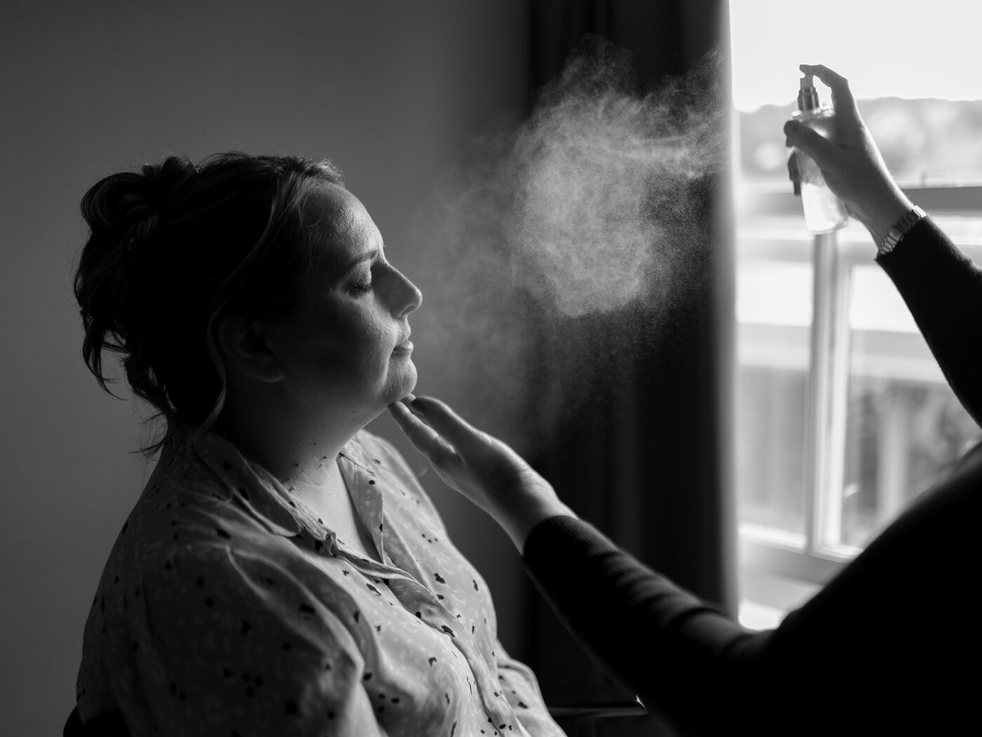 Black-and-white photo of a bridesmaid having makeup applied near a window as light and mist highlight her profile.
