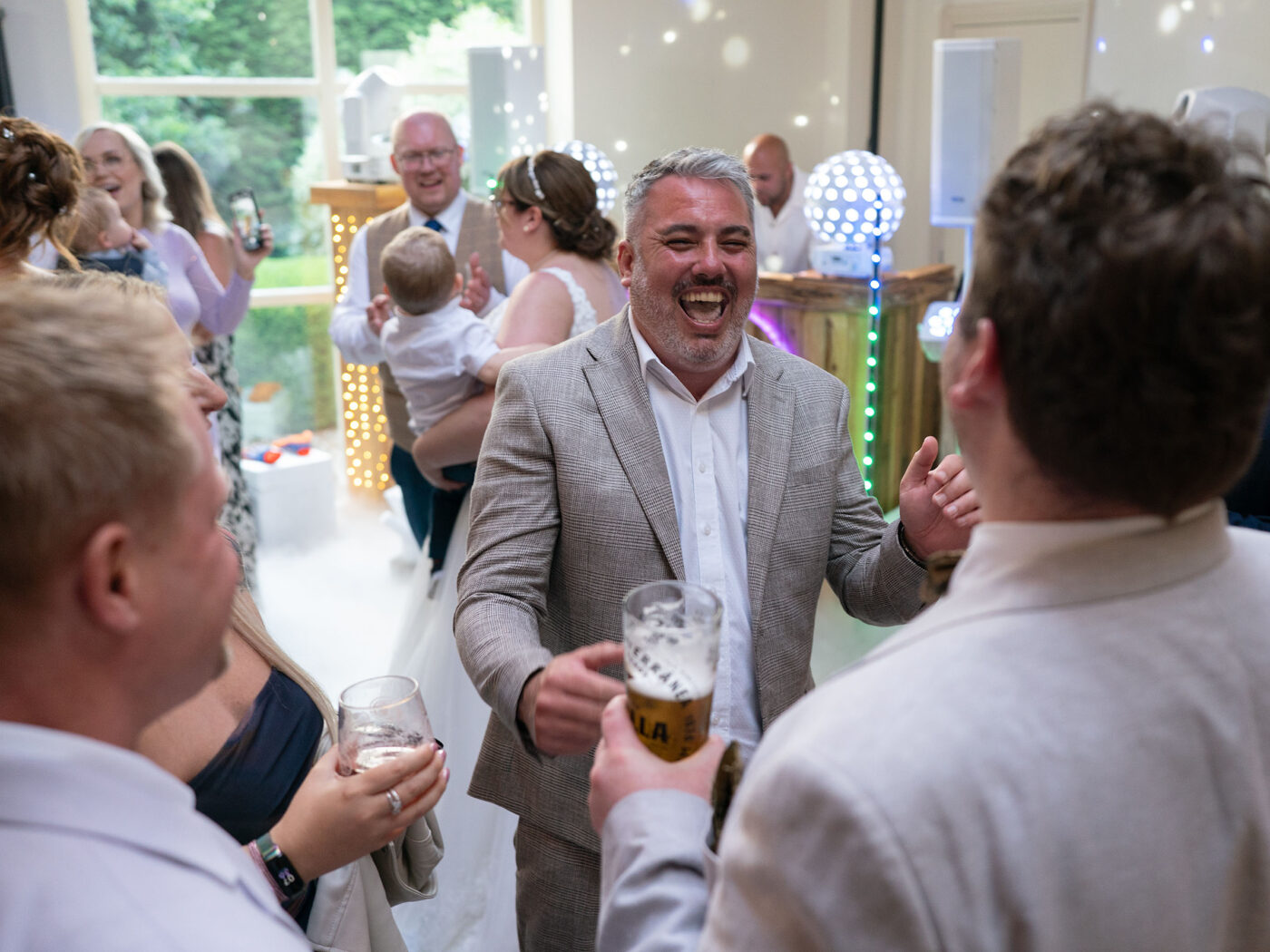 Guests chat and laugh indoors during a wedding reception while holding drinks in a bright party setting at Shottle Hall