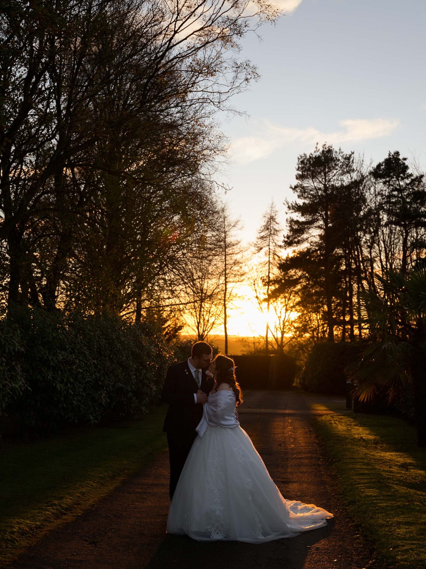Bride and groom kiss on a tree-lined path at sunset, silhouetted by warm golden light at Goosedale