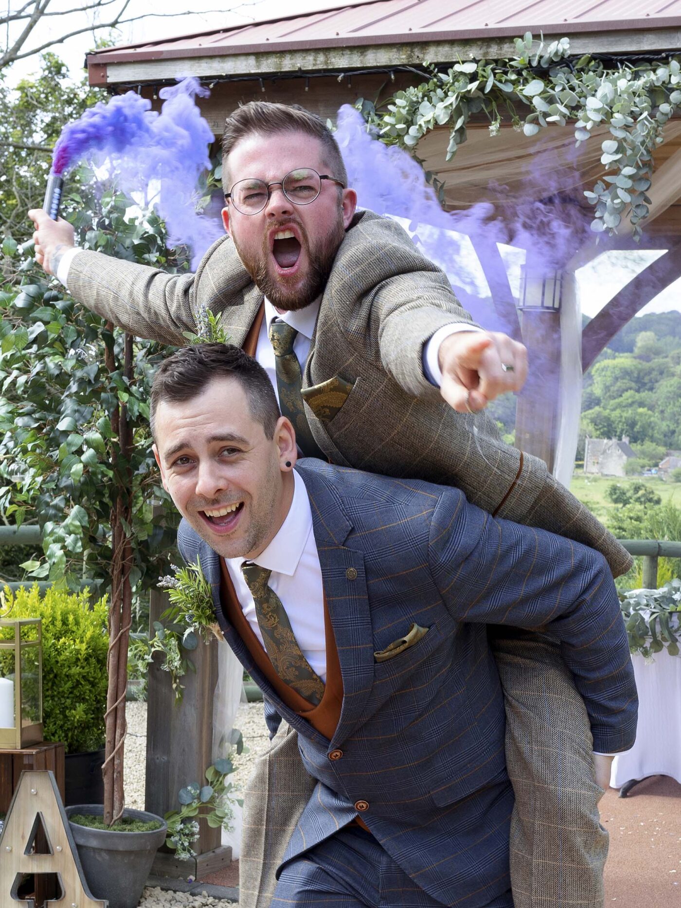 Two suited Grooms pose playfully, one giving the other a piggyback ride while pointing toward the camera.