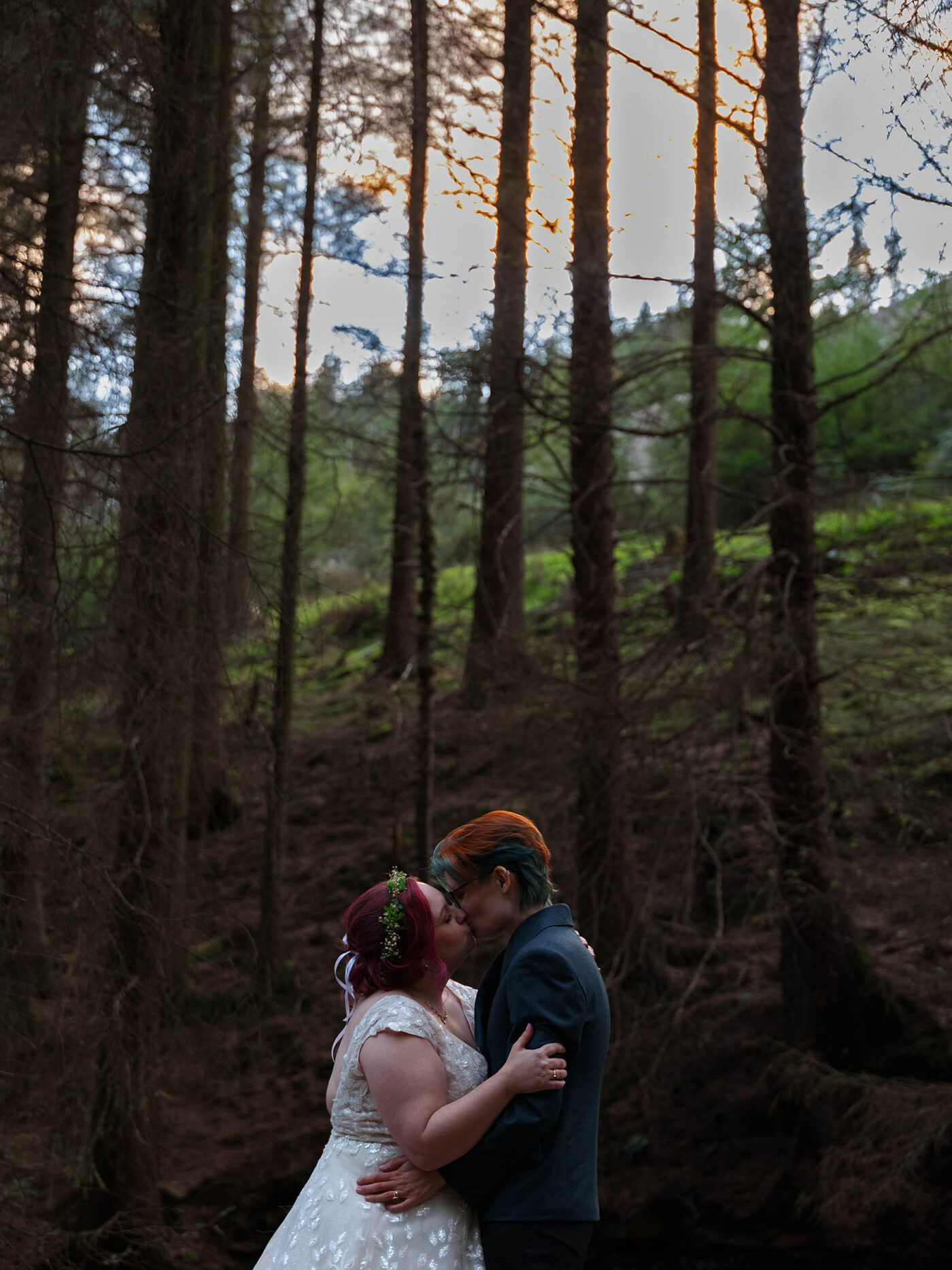 Newlywed couple share a kiss in a peak district woodland sunset, surrounded by tall trees and warm evening light.